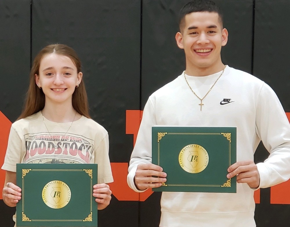 teen girl with long, light brown hair wearing a tan Woodstock t-shirt and a young man with very short  black hair wearing a white sweatshirt with a gold cross necklace holding green and gold certificates